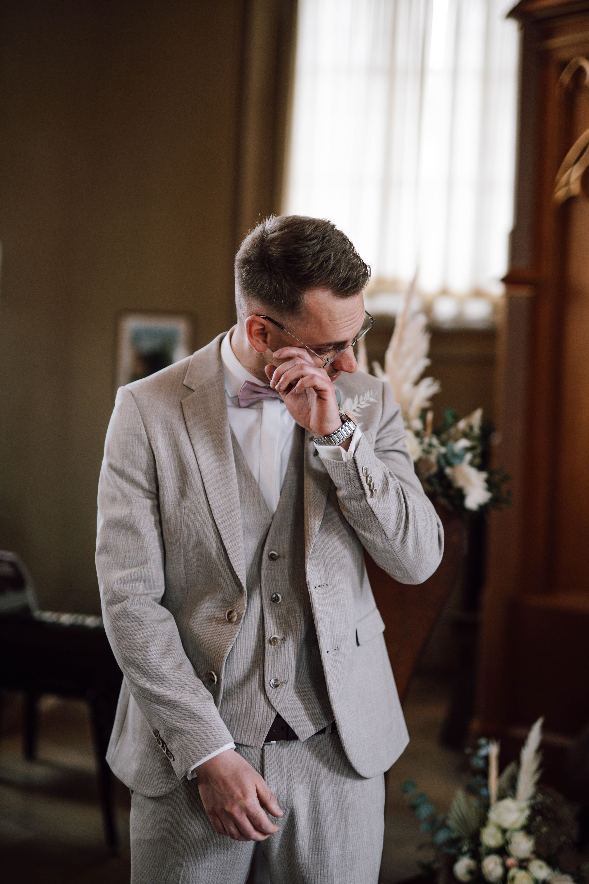 Emotional groom in light gray suit wiping tears during wedding ceremony.