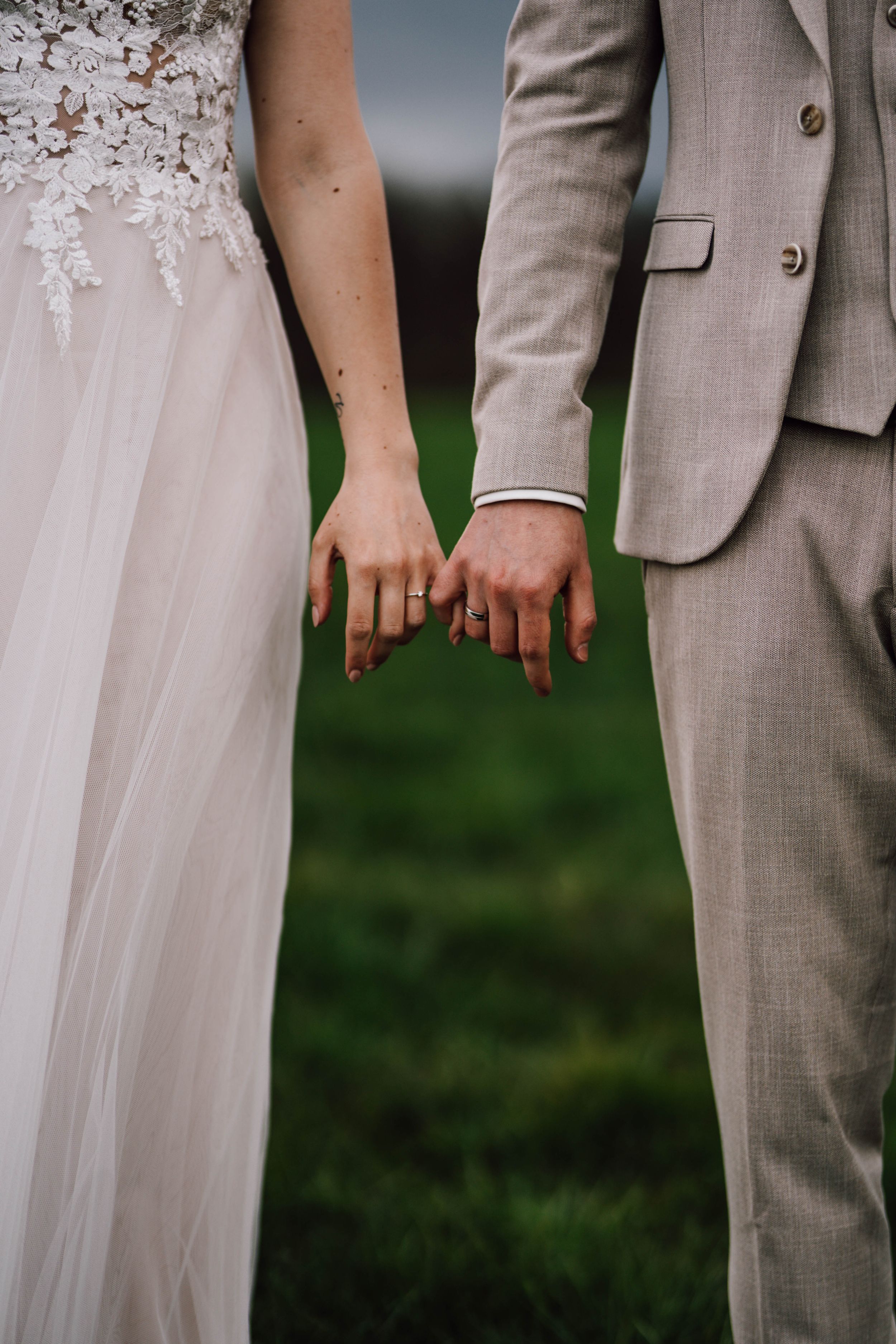 Close-up of newlyweds holding hands, bride in lace dress and groom in light gray suit against green background.