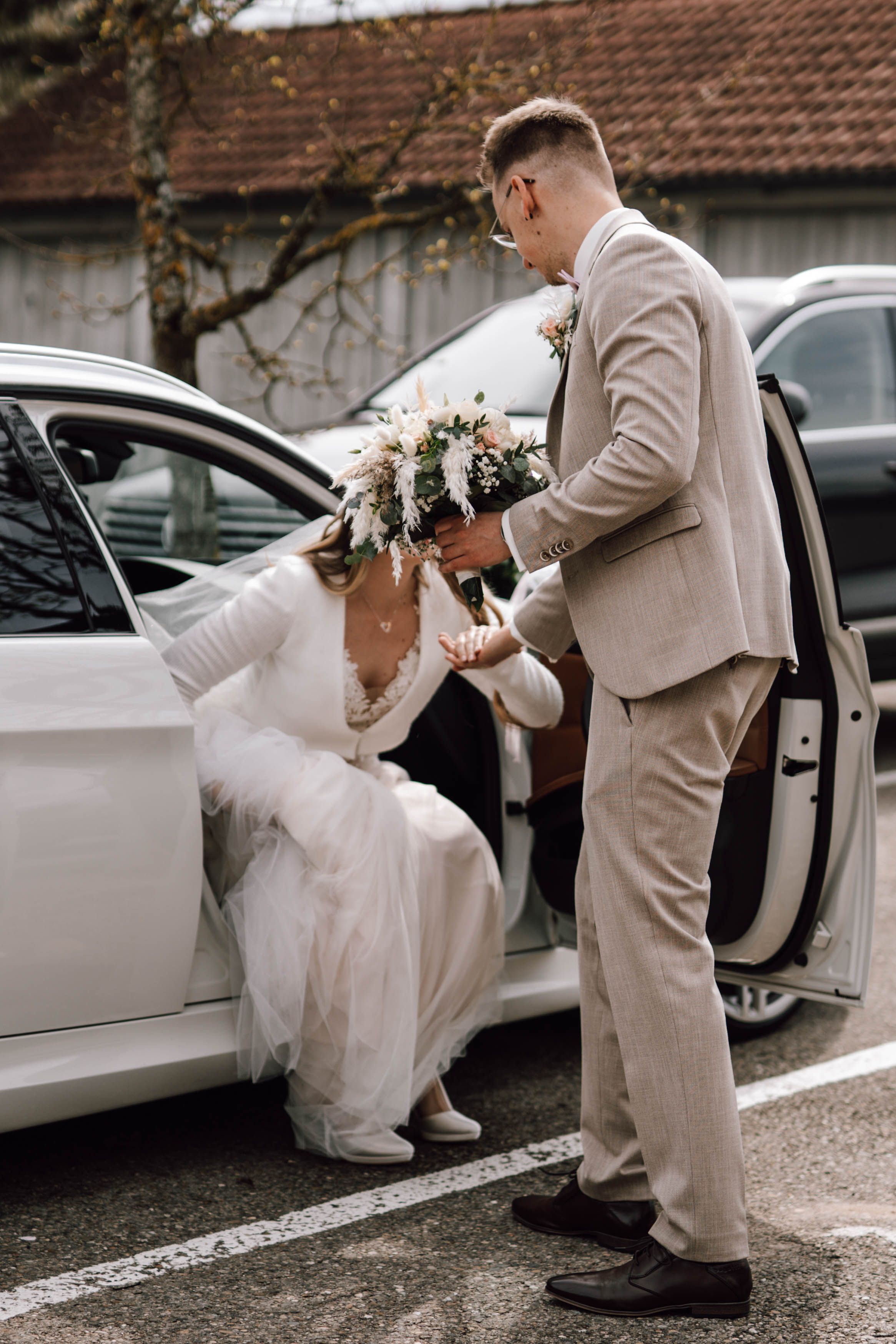 Groom in beige suit helping bride into white car with wedding bouquet.
