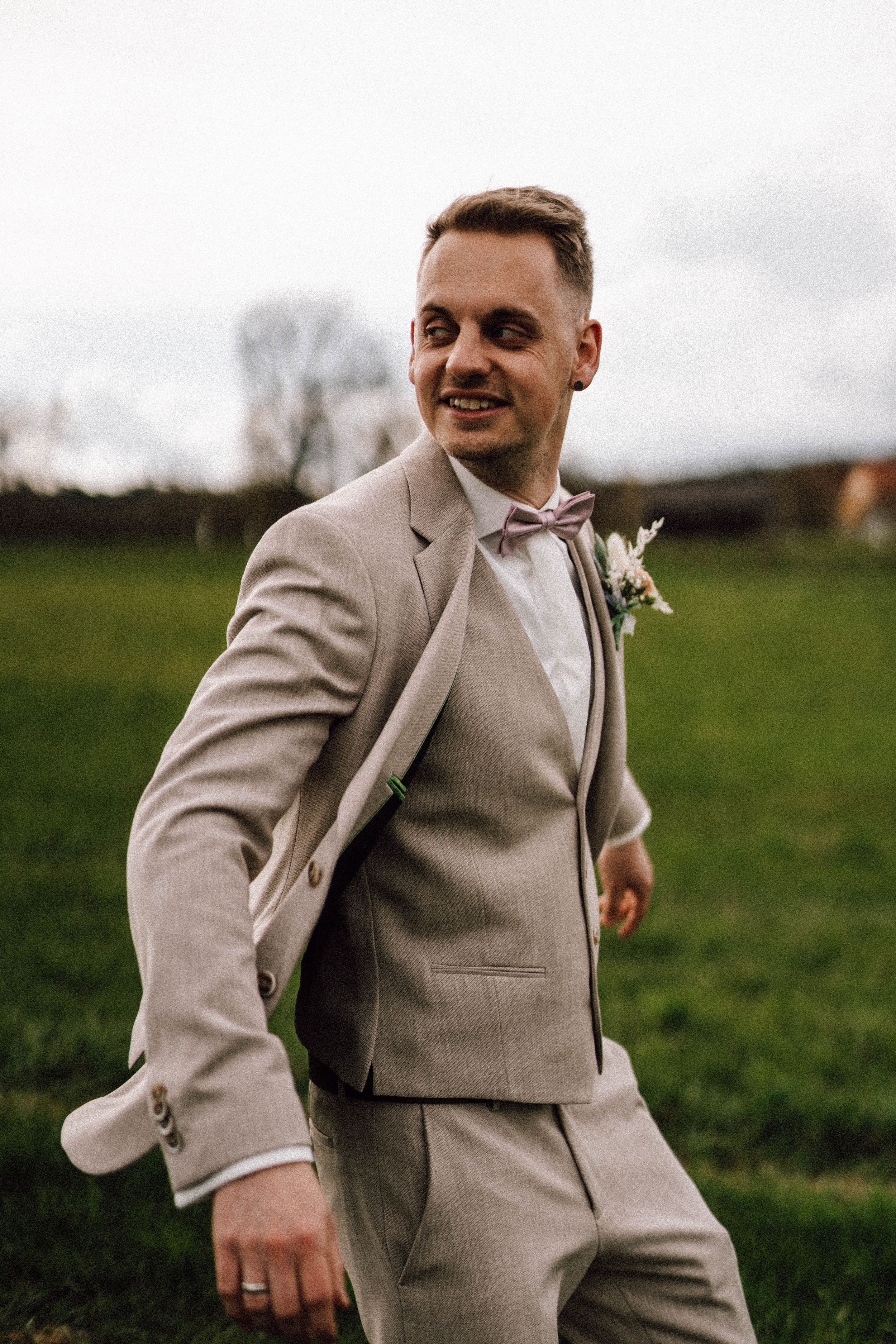 Groom in beige suit walking joyfully across green field.