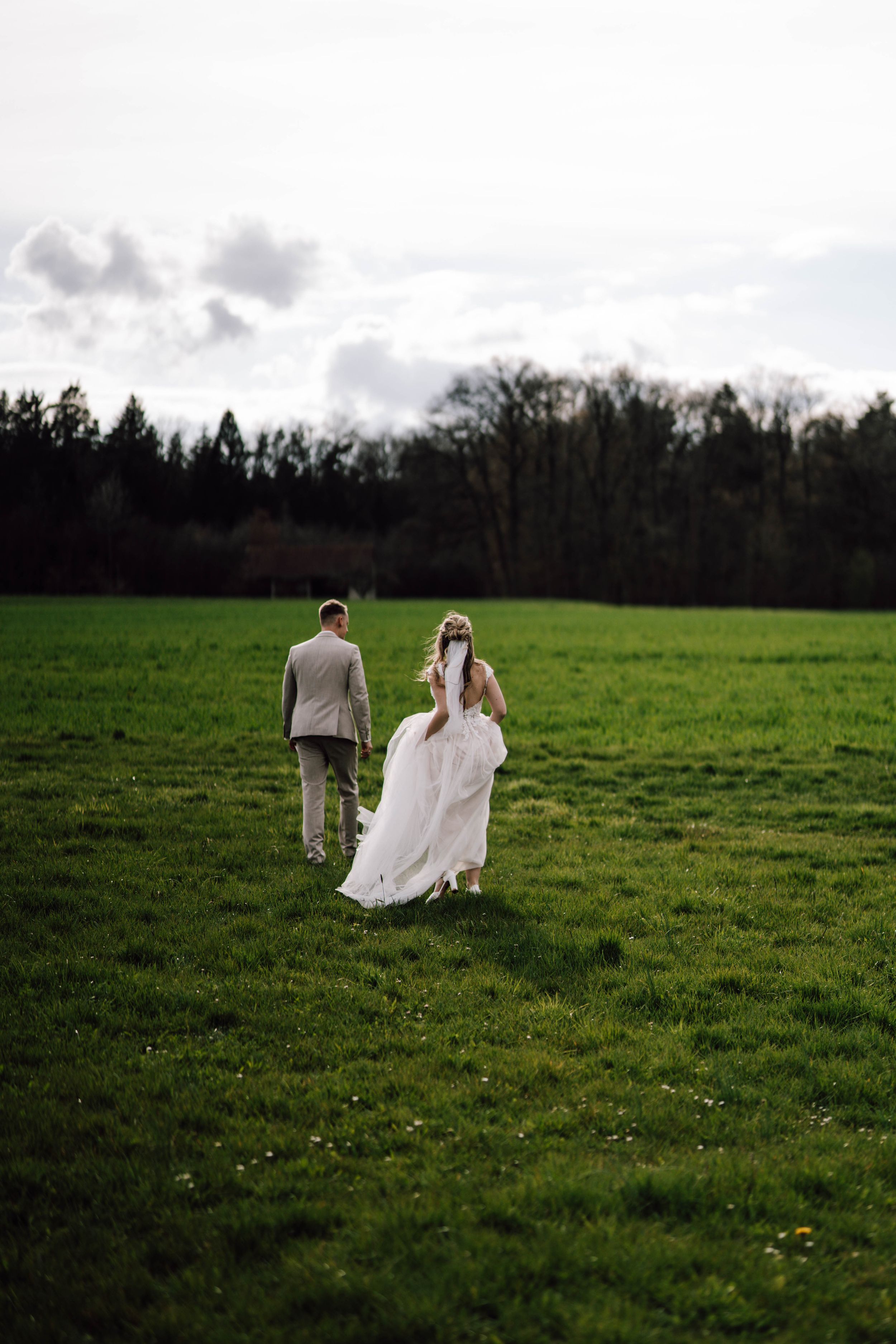 A couple walks through a green field with trees in the background on their wedding day.