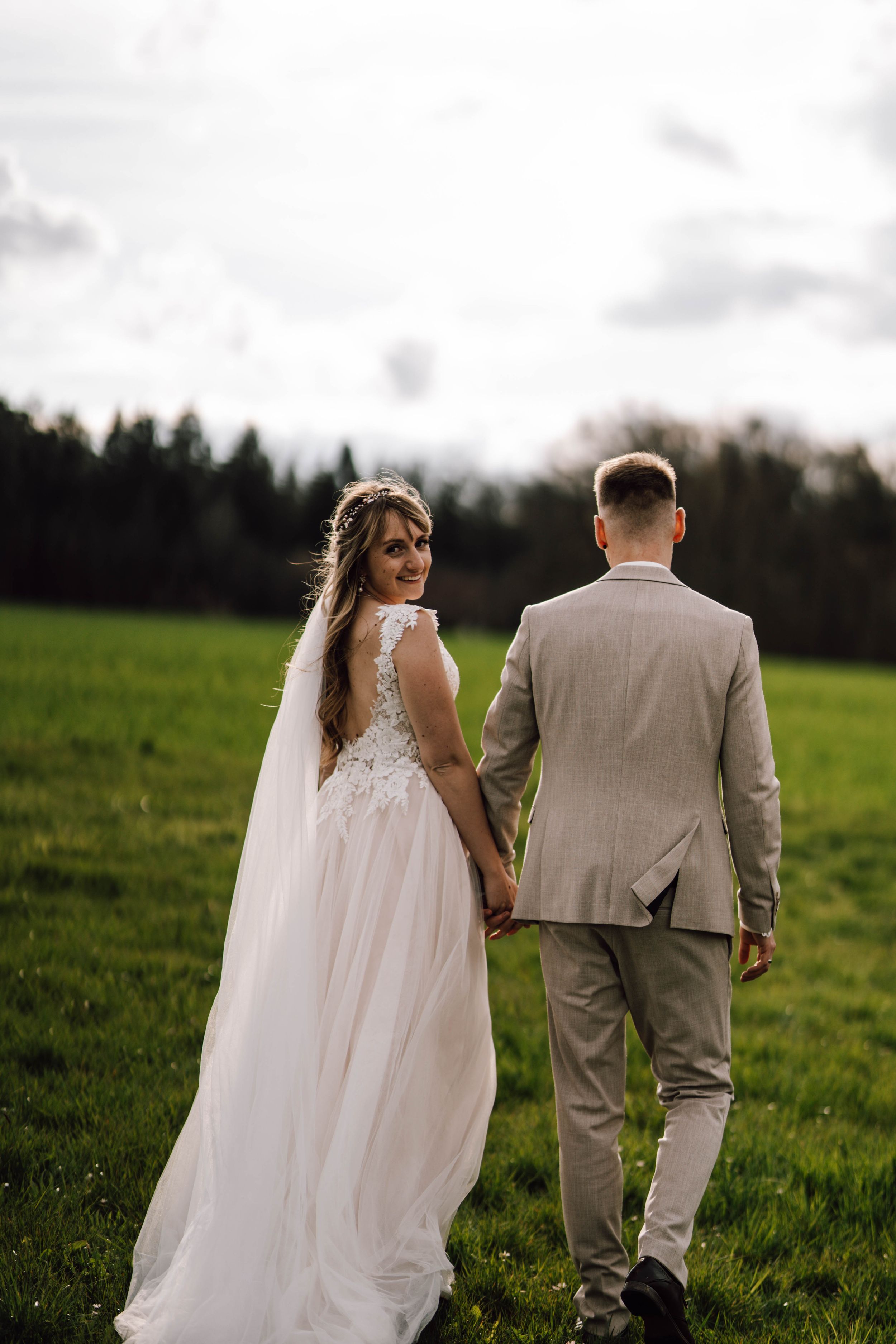 A couple walks hand in hand through a grassy field in wedding attire.