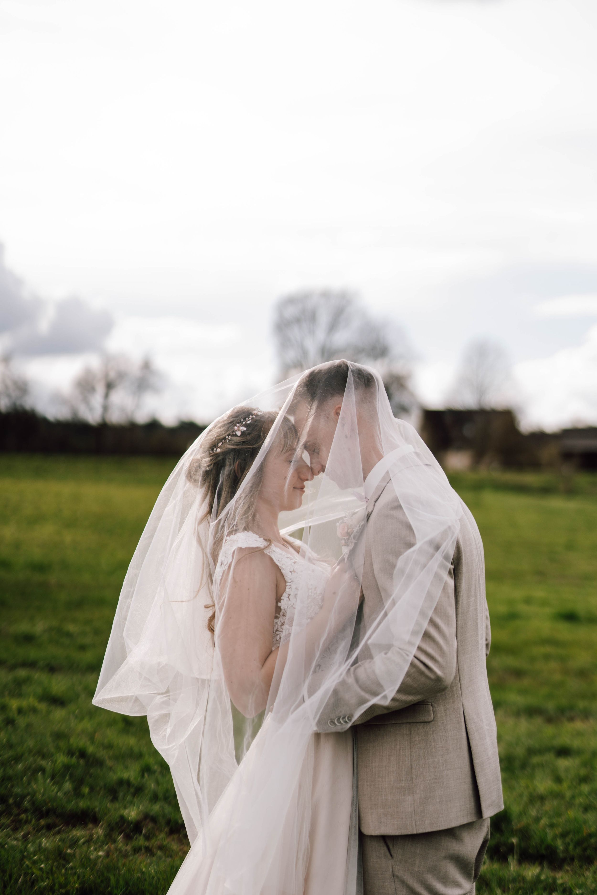Couple embraces under flowing white veil in outdoor wedding setting.