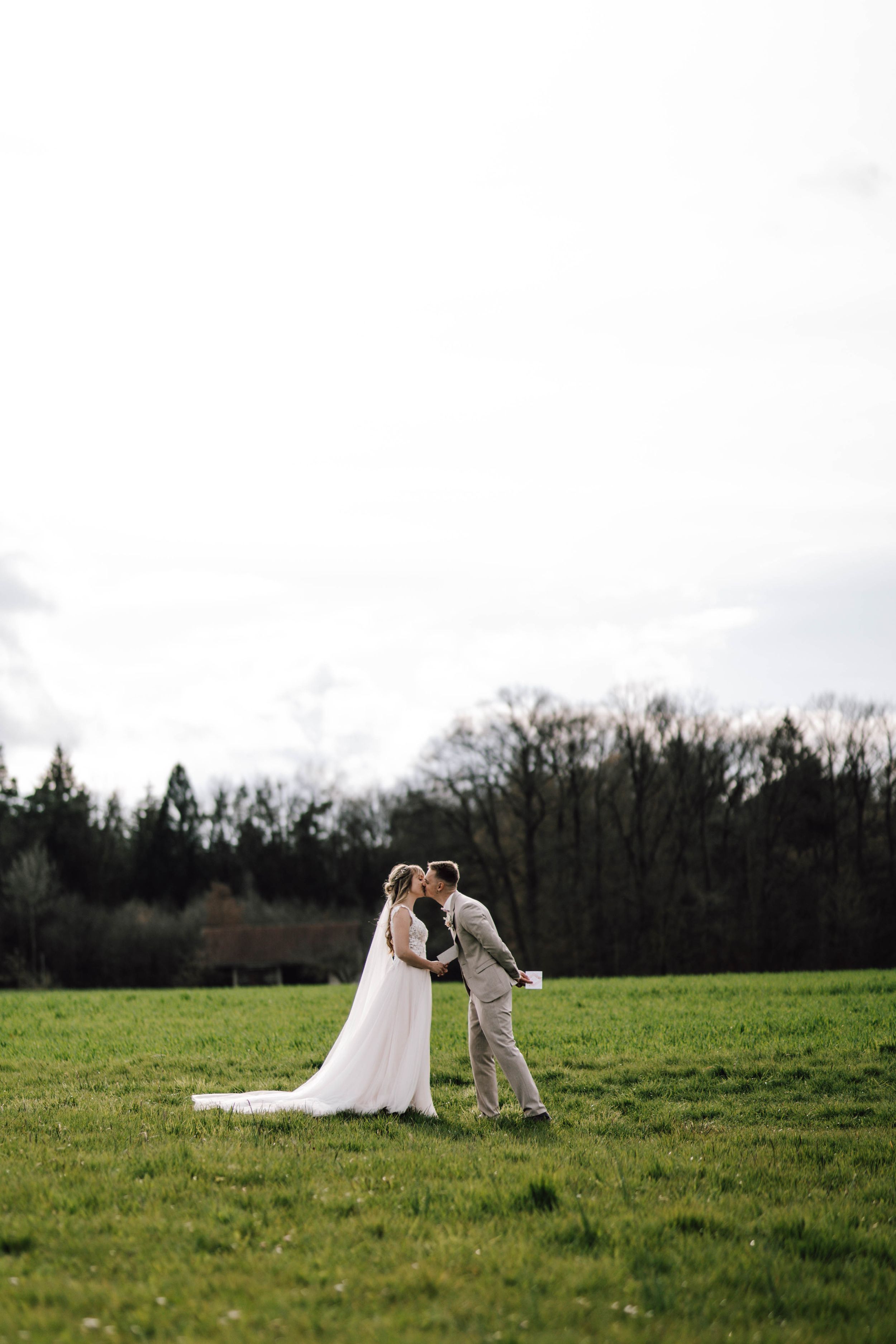 Newlyweds share a romantic moment in a sunlit meadow surrounded by trees.