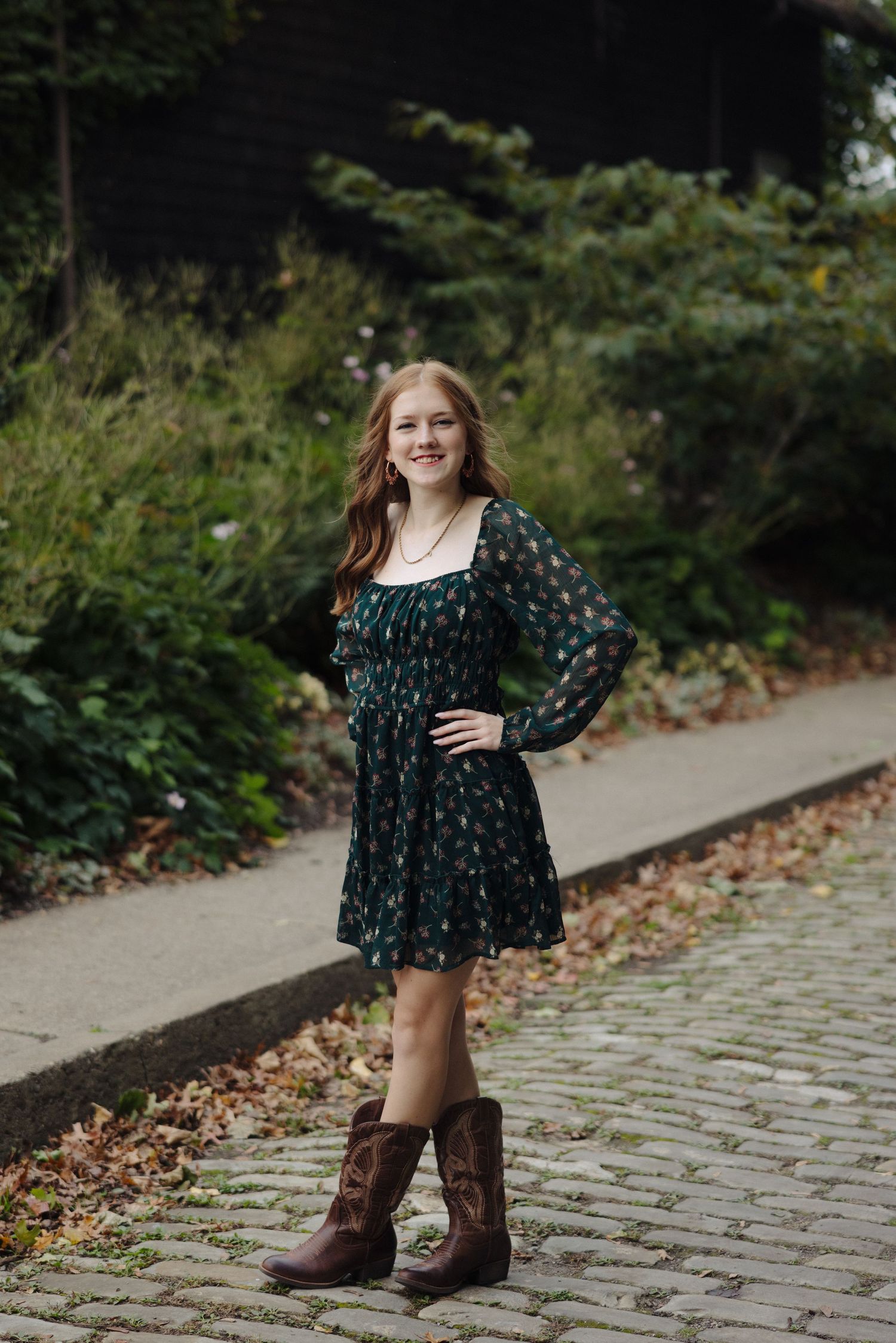 A person in a floral dress and brown boots poses on a cobblestone path.
