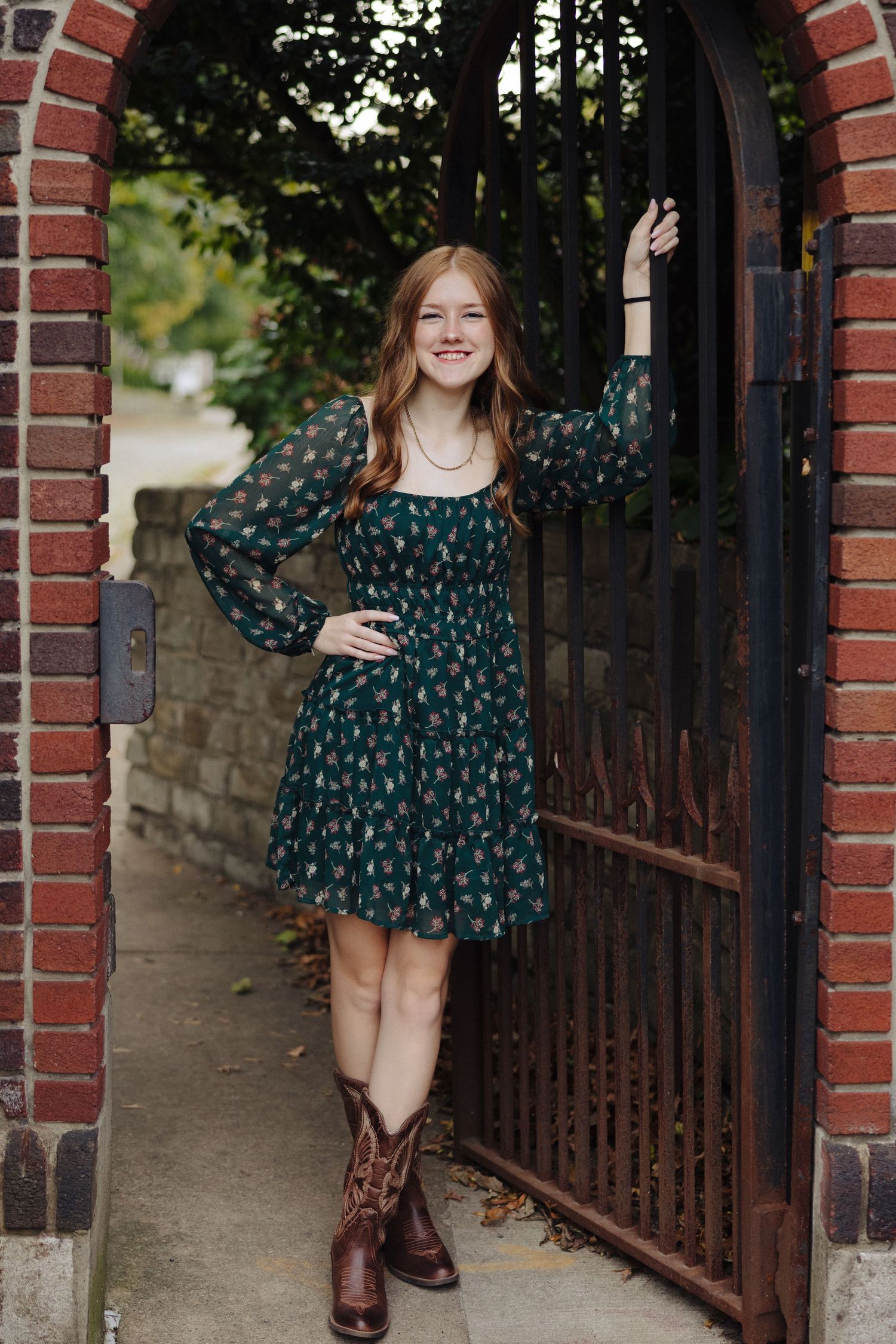 A person in a floral dress and boots poses by a brick archway with an iron gate.