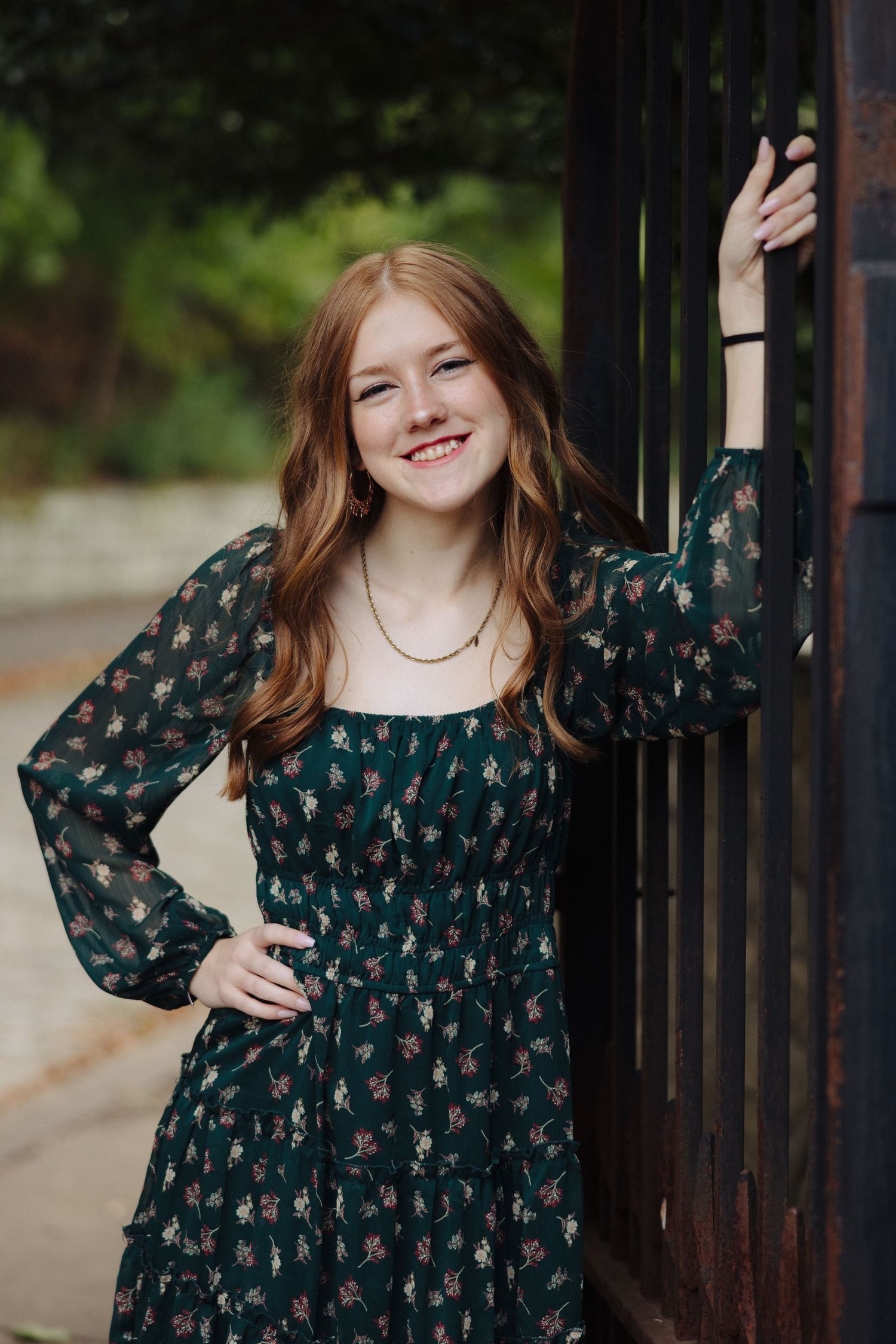 A person in a dark floral dress poses against an iron gate with a cheerful expression.