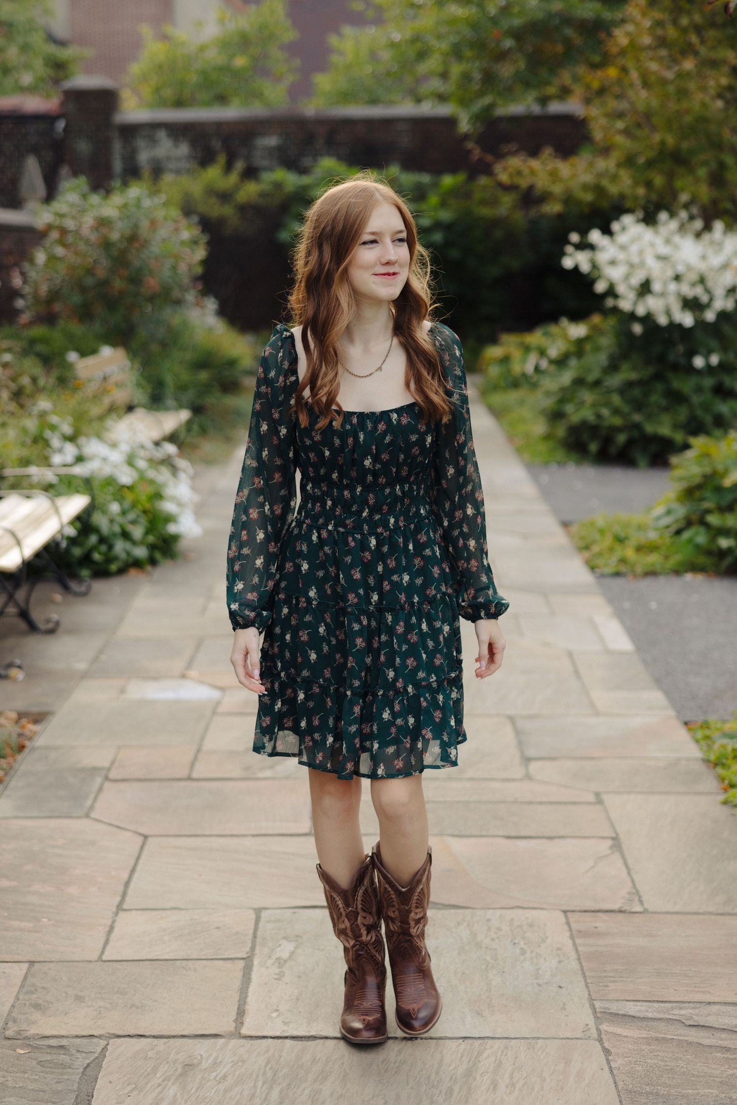 Woman in dark floral dress and brown cowboy boots stands on stone pathway in garden setting.