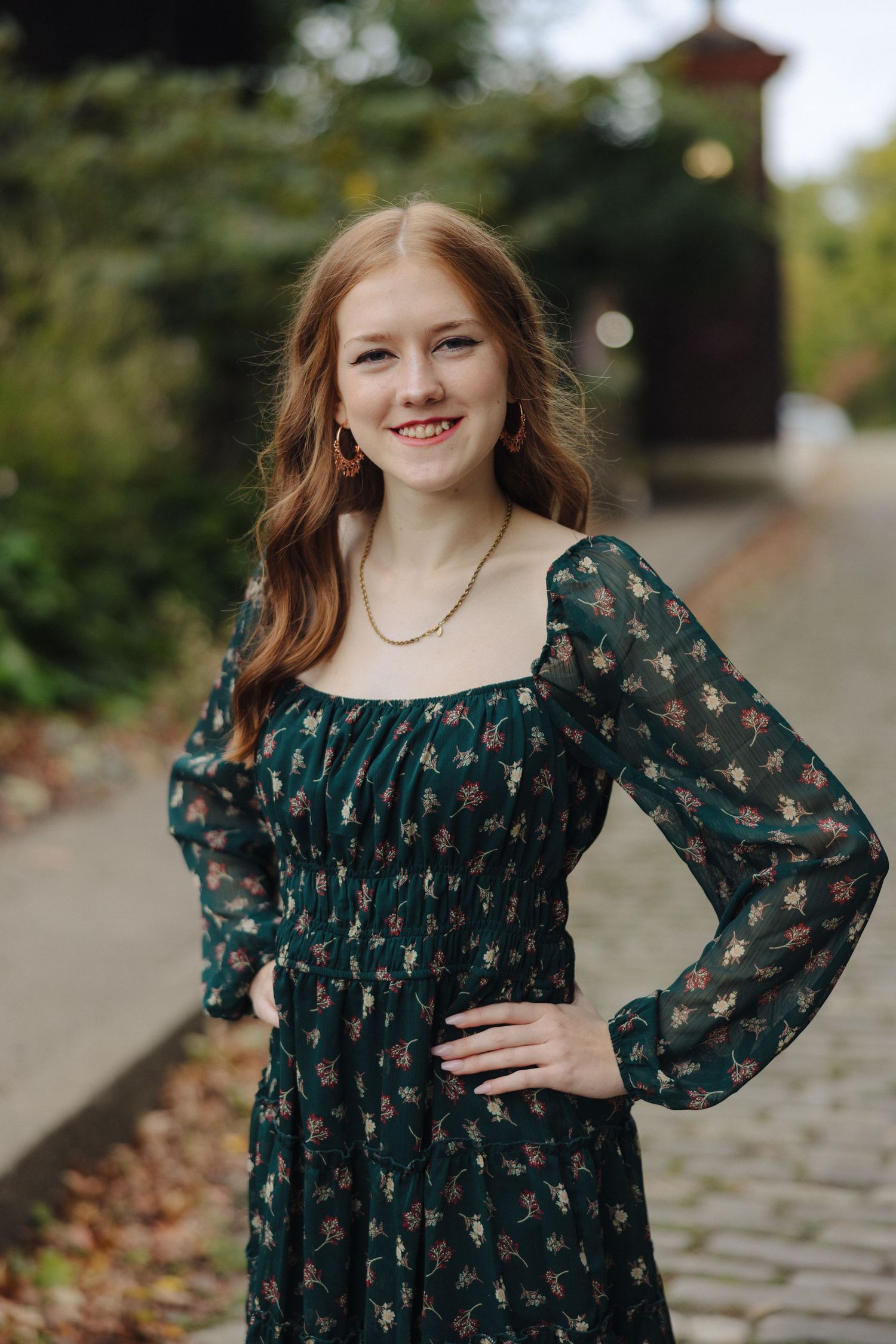 A smiling woman in a dark green patterned dress with puff sleeves poses in front of a garden setting.