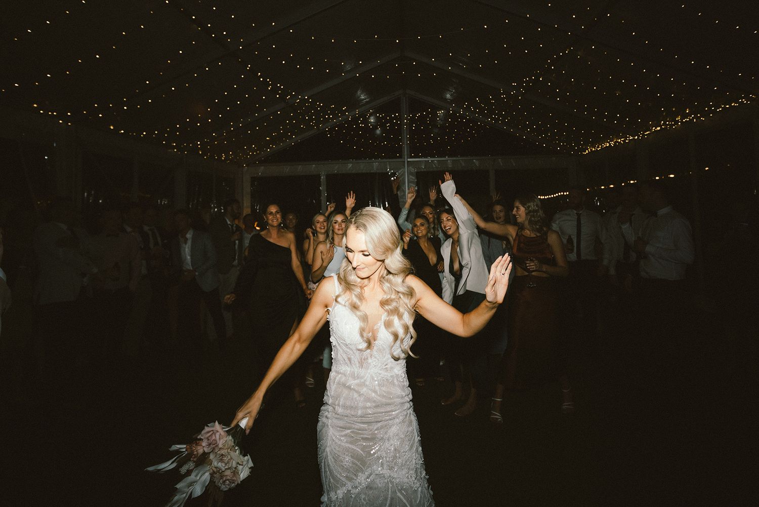 Wedding reception dance floor filled with guests celebrating under romantic string lighting.