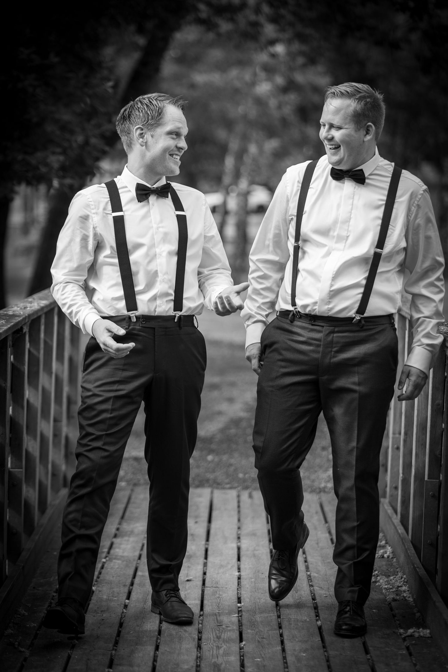Two groomsmen in black and white attire with suspenders laughing on wooden bridge.