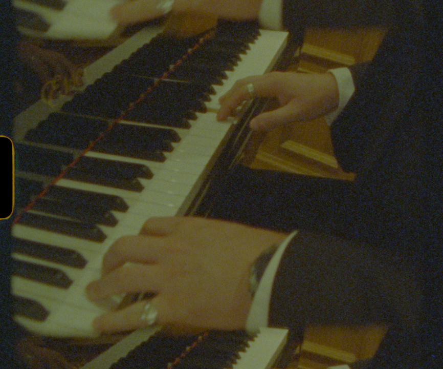 Hands of two pianists playing keys on a grand piano in a dimly lit concert setting.