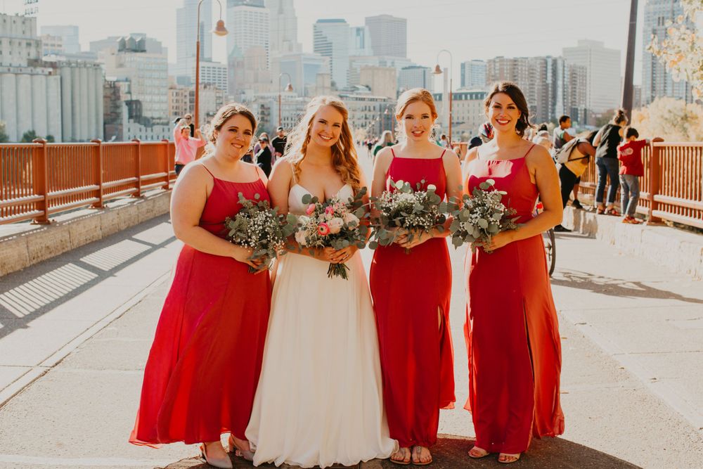 Smiling bride and three bridesmaids in red gowns hold greenery bouquets on a sunny urban bridge.