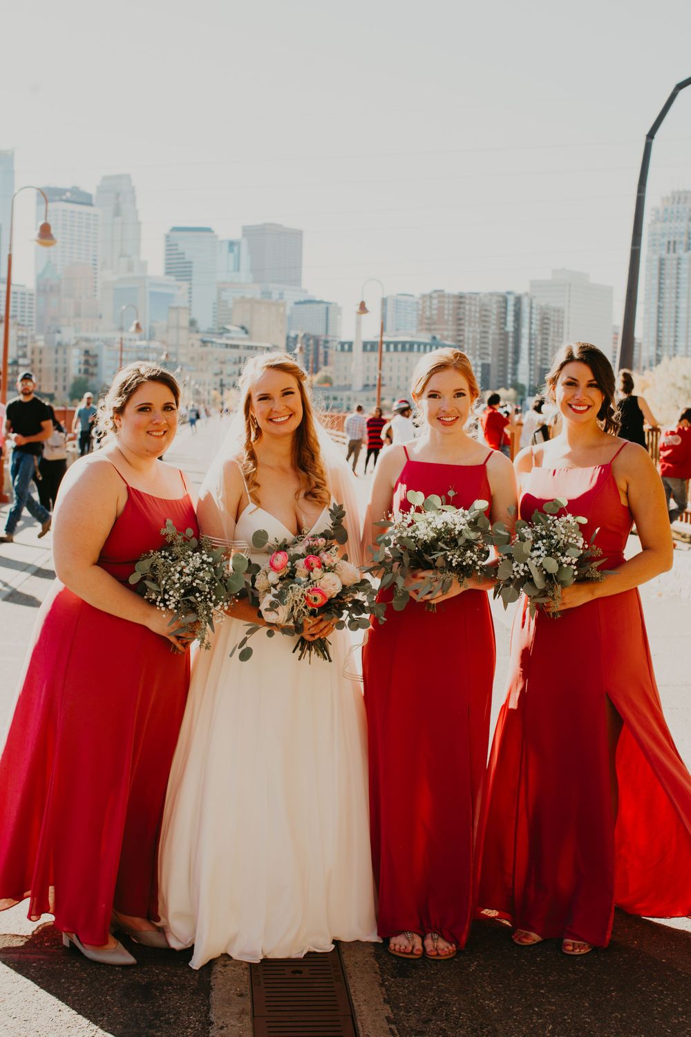 Bride in white gown poses with three bridesmaids in red dresses holding bouquets with city skyline backdrop.
