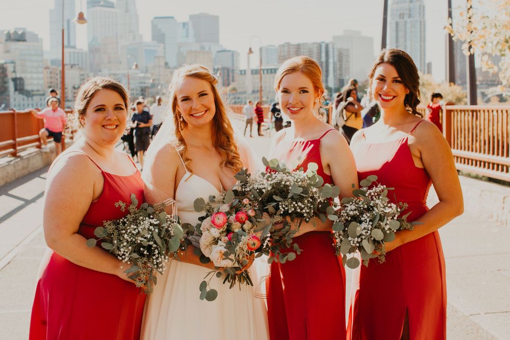 Bride and three bridesmaids in red dresses smile holding lush green and pink bouquets outdoors.