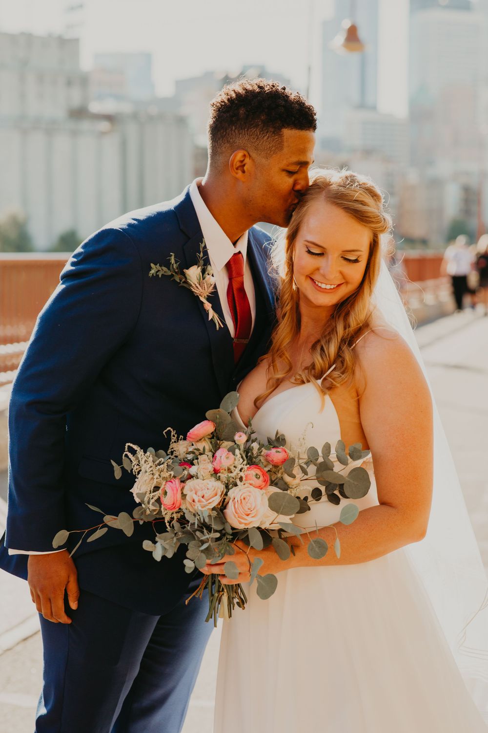 Groom in navy suit kisses bride on forehead as she smiles holding a pink and blush bouquet.