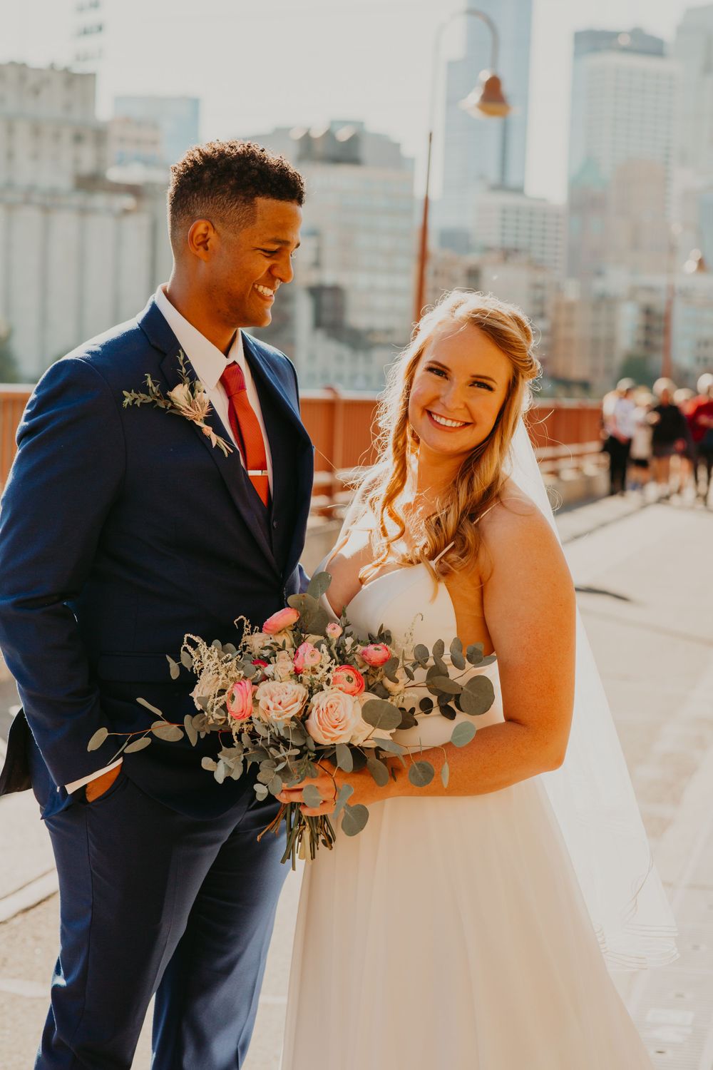 Bride smiles beside groom in navy suit holding a pink rose and eucalyptus bouquet outdoors.