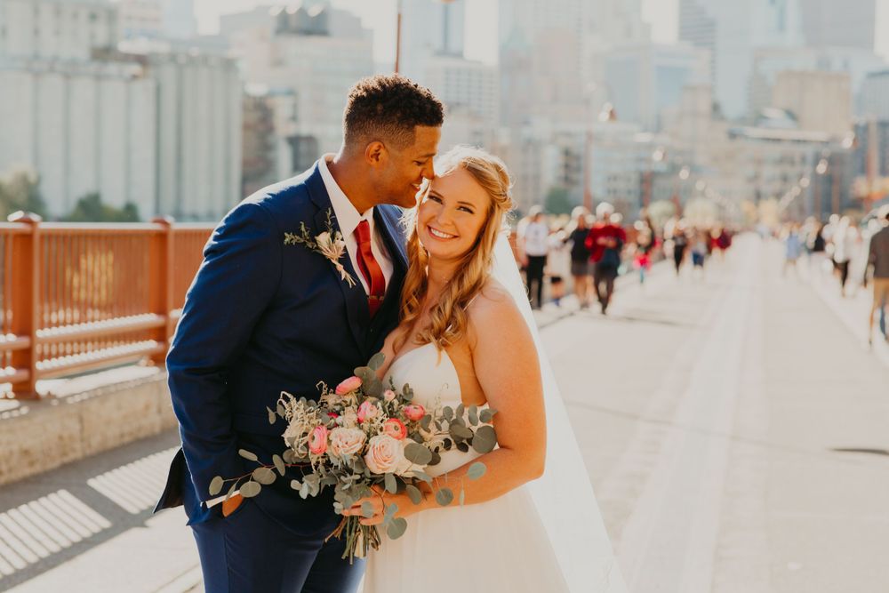 Groom in navy suit and red tie laughs with smiling bride holding bouquet on a sunny bridge.