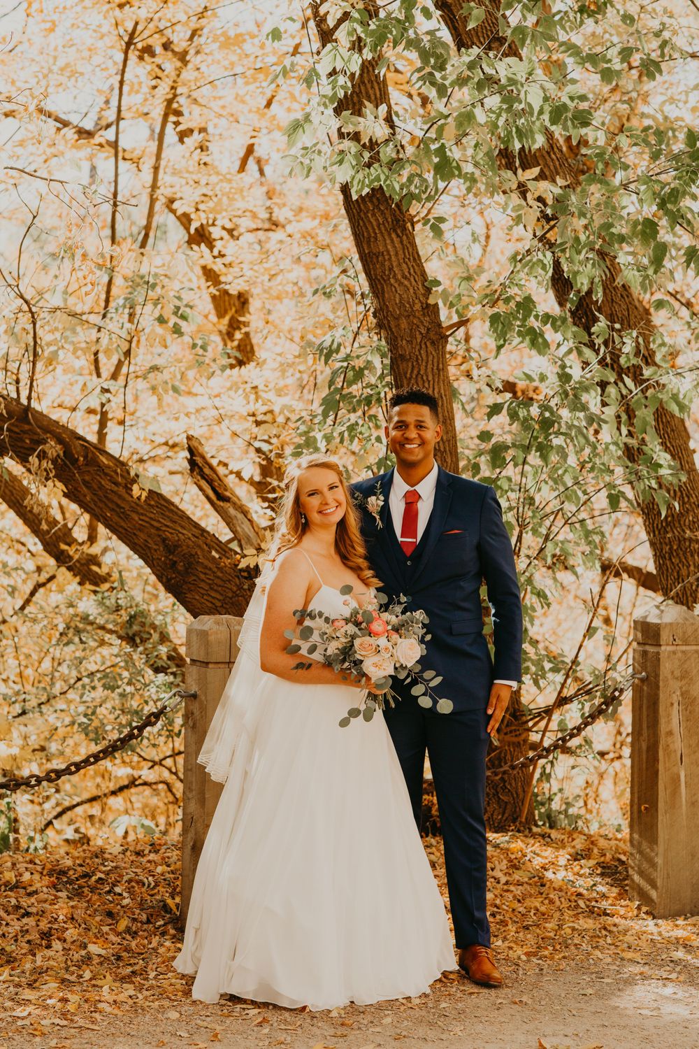 Bride and groom smile together under golden autumn trees holding a blush and eucalyptus bouquet.