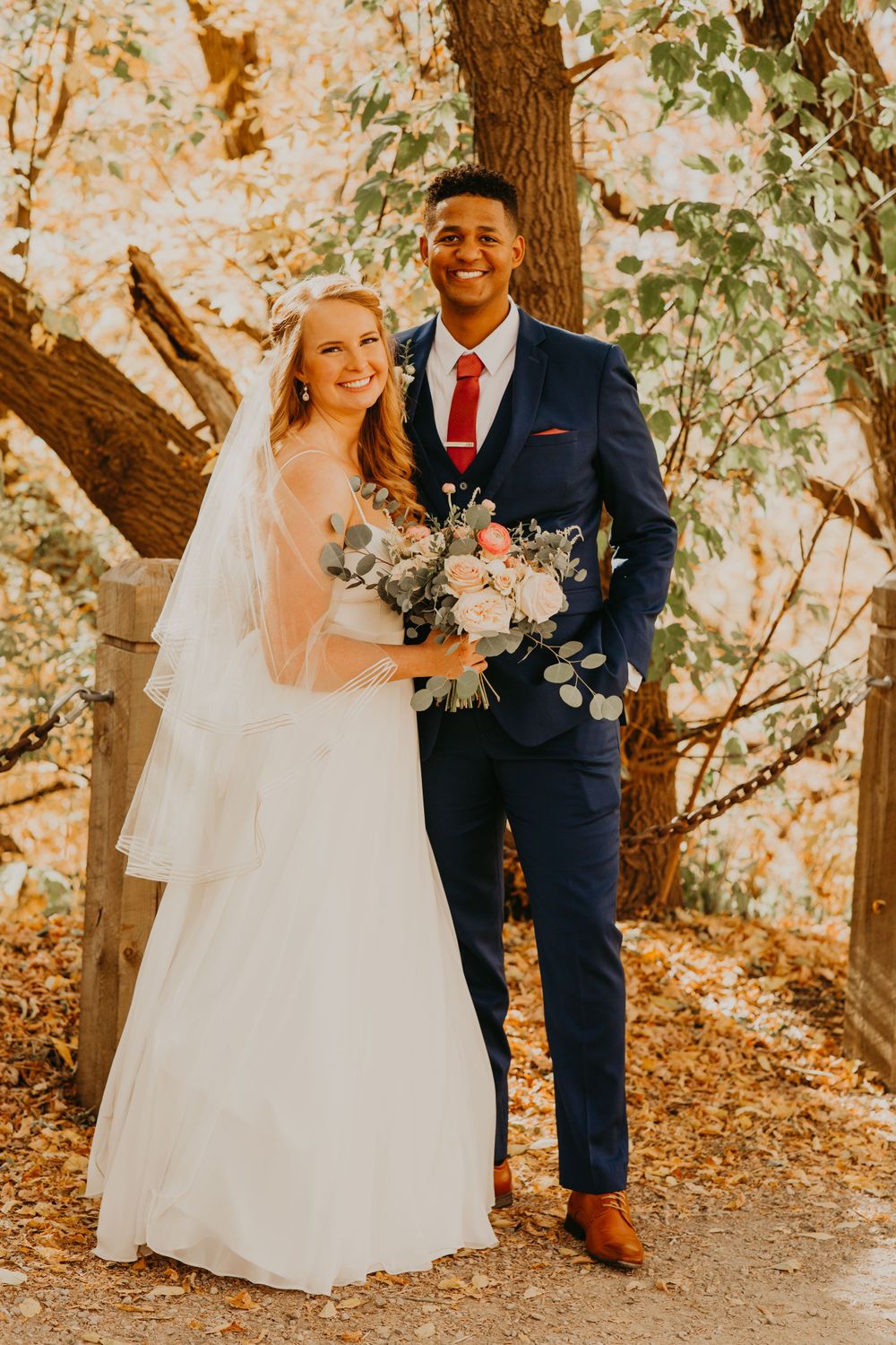 Smiling interracial wedding couple posing outdoors in fall foliage, bride in white gown with veil holding bouquet, groom in navy suit.
