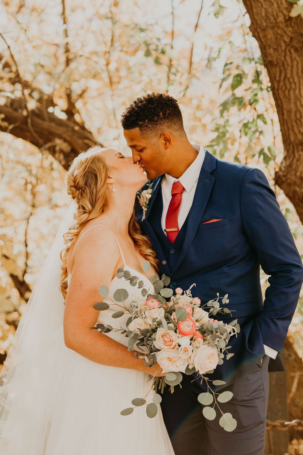 Interracial newlyweds sharing a kiss outdoors in autumn, groom in navy suit with red tie, bride holding pink and white bouquet.