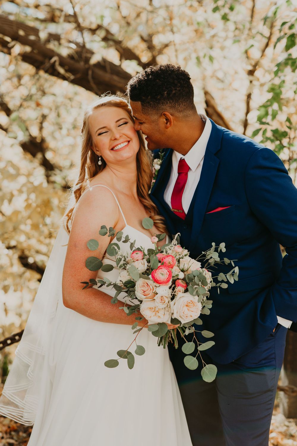 Groom in navy suit kissing bride on cheek as she smiles, bride holding pink and white rose bouquet in autumn setting.