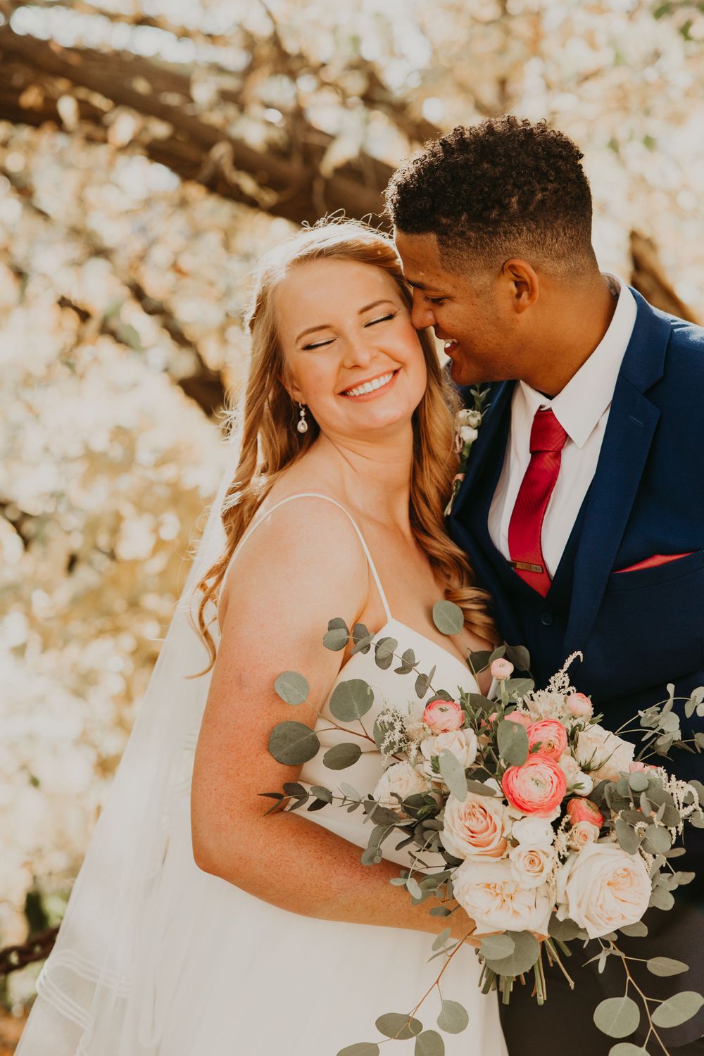 Joyful bride smiling as groom whispers in her ear, holding floral bouquet with pink roses and eucalyptus in warm fall light.