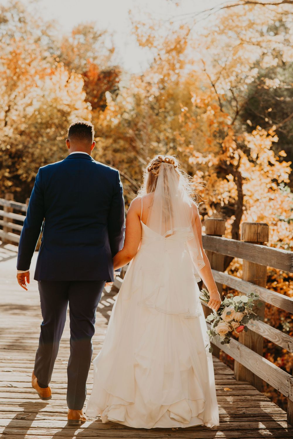 Bride and groom walking hand in hand on wooden bridge surrounded by vibrant fall foliage, viewed from behind.
