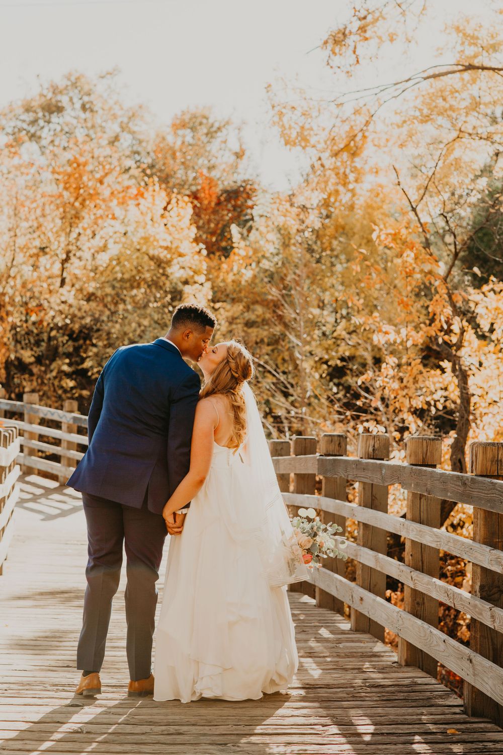 Newlyweds sharing a romantic kiss on a wooden bridge surrounded by golden autumn trees on their wedding day.