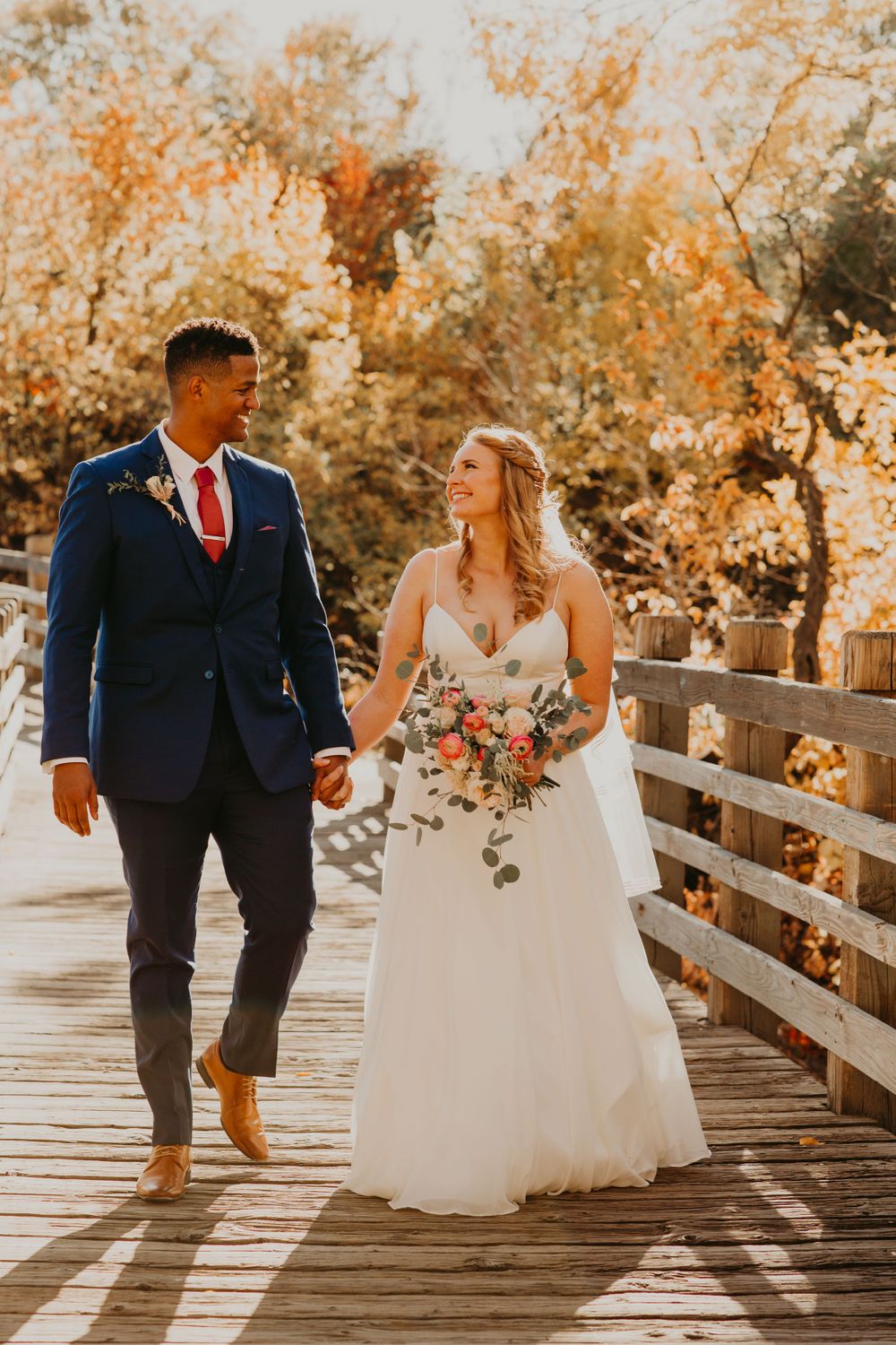 Happy interracial couple walking hand in hand on wooden bridge, bride in white gown holding bouquet, groom in navy suit.