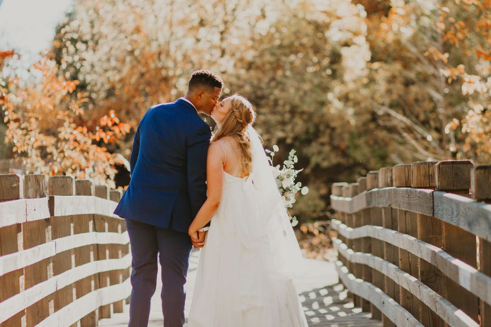 Bride and groom kissing on a rustic wooden bridge surrounded by soft golden autumn foliage on their wedding day.
