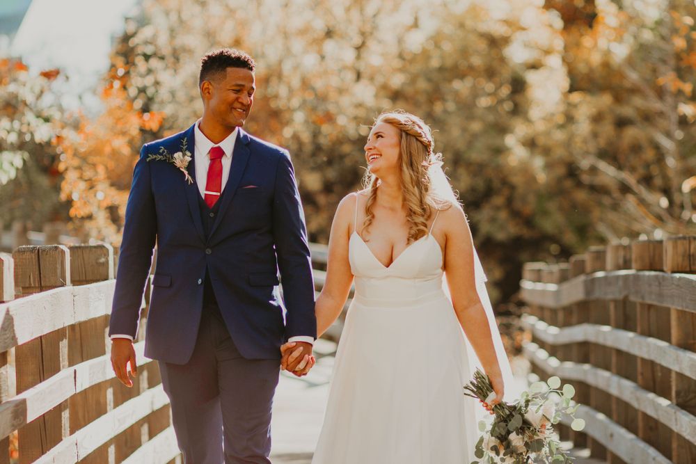 Smiling interracial newlyweds walking hand in hand on a wooden bridge with warm golden fall foliage in the background.