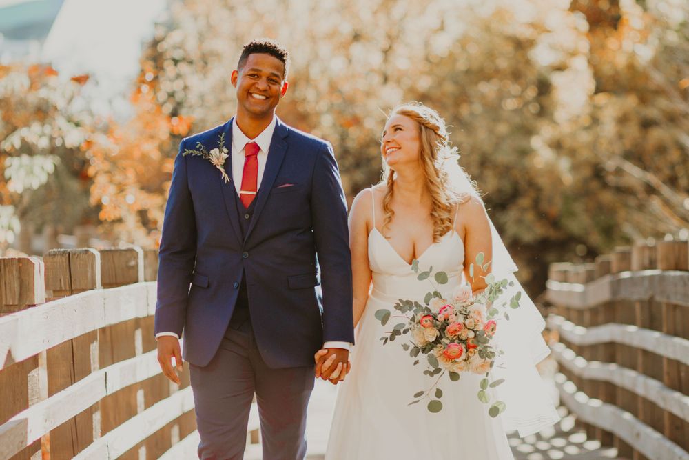 Interracial wedding couple strolling on wooden bridge, bride in white gown with pink bouquet, groom in navy suit and red tie.