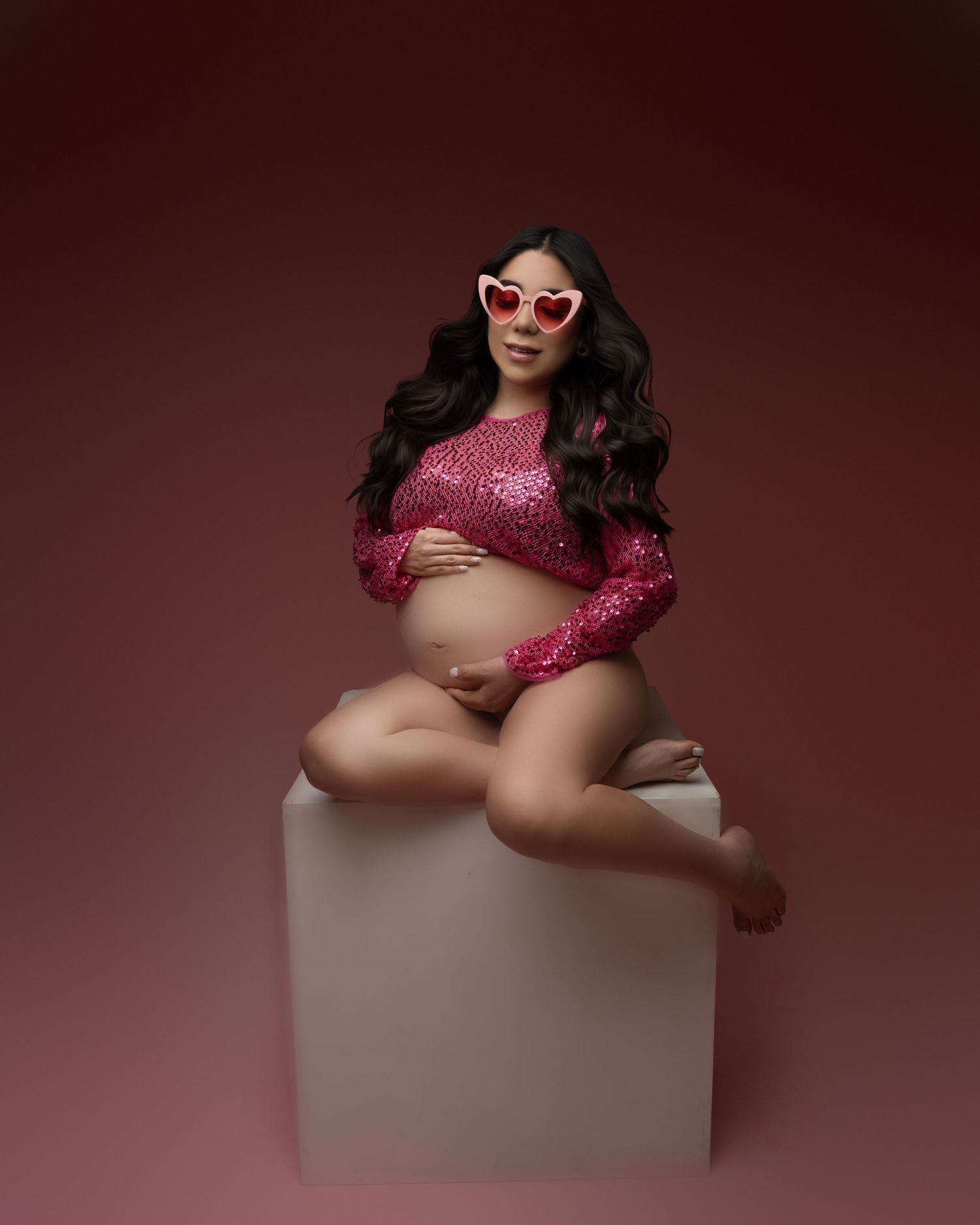 Stylish maternity boudoir photo with pink sequin top and white sunglasses on neutral backdrop.
