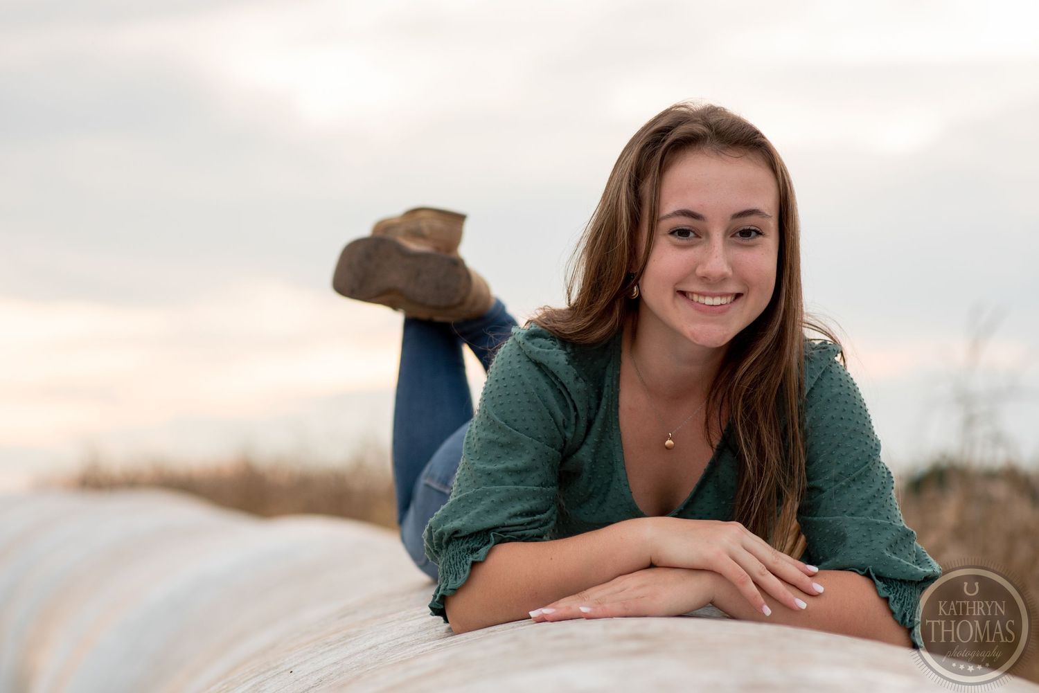 Relaxed outdoor portrait of someone lying on a hay bale wearing jeans and a green top at dusk.