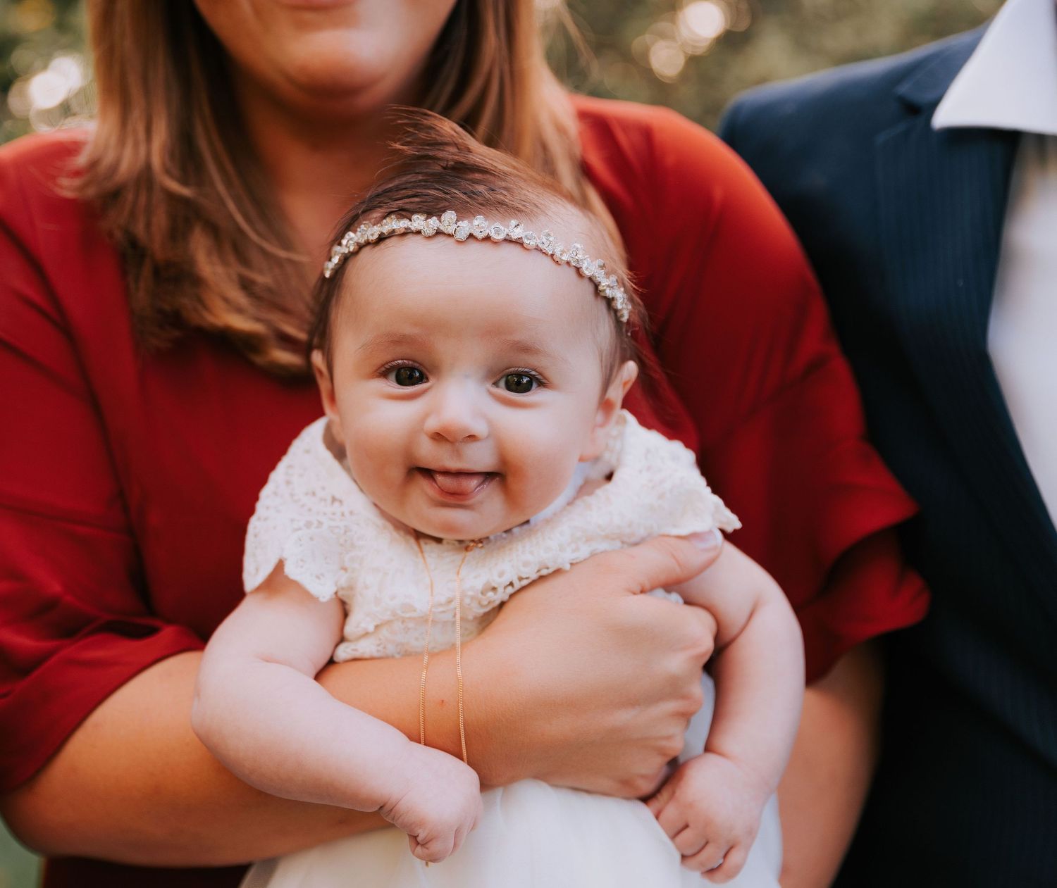 A baby wearing a white lace dress and headband is held during a baptism ceremony.