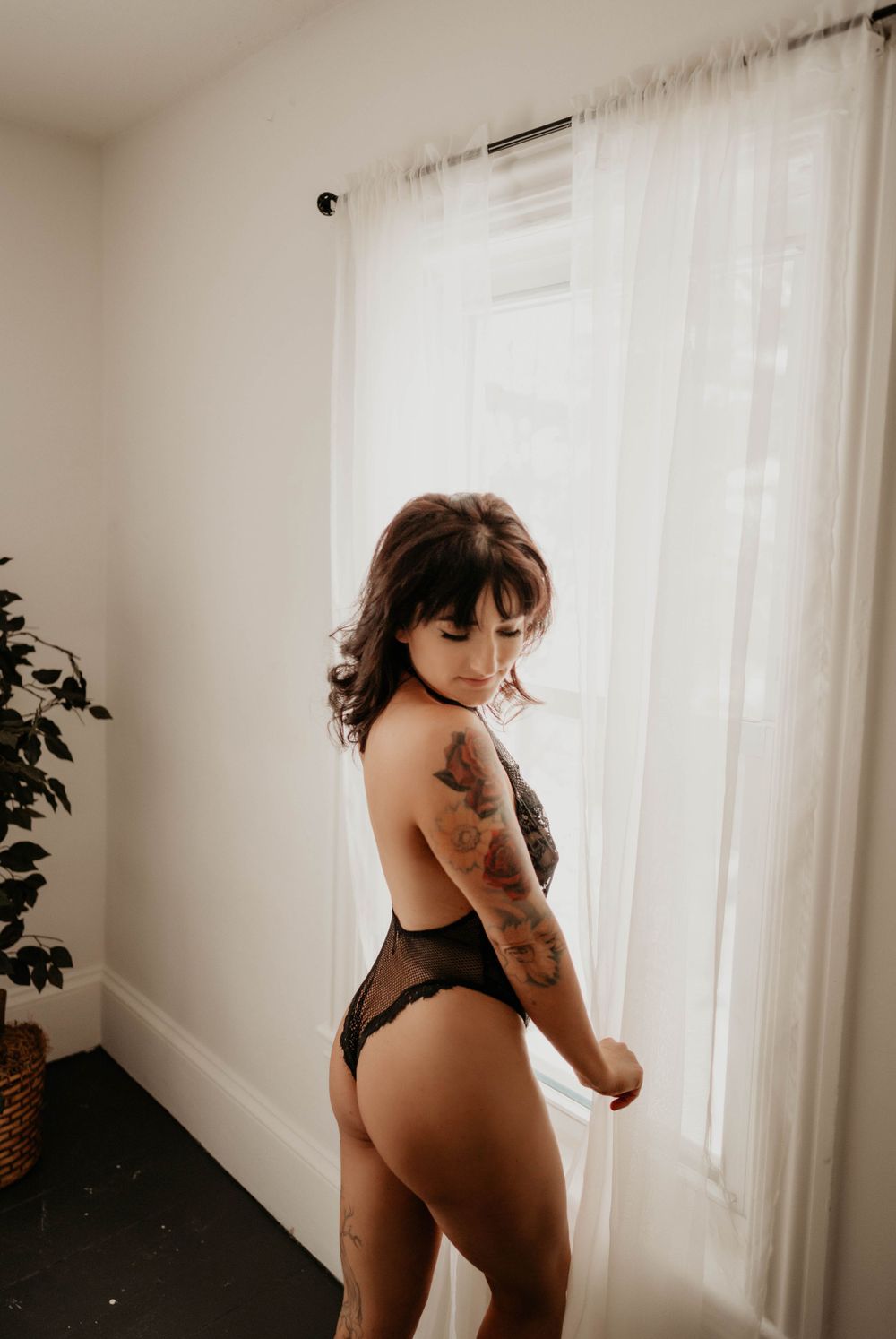 Person in black lingerie with tattoos posing near sheer curtained window.