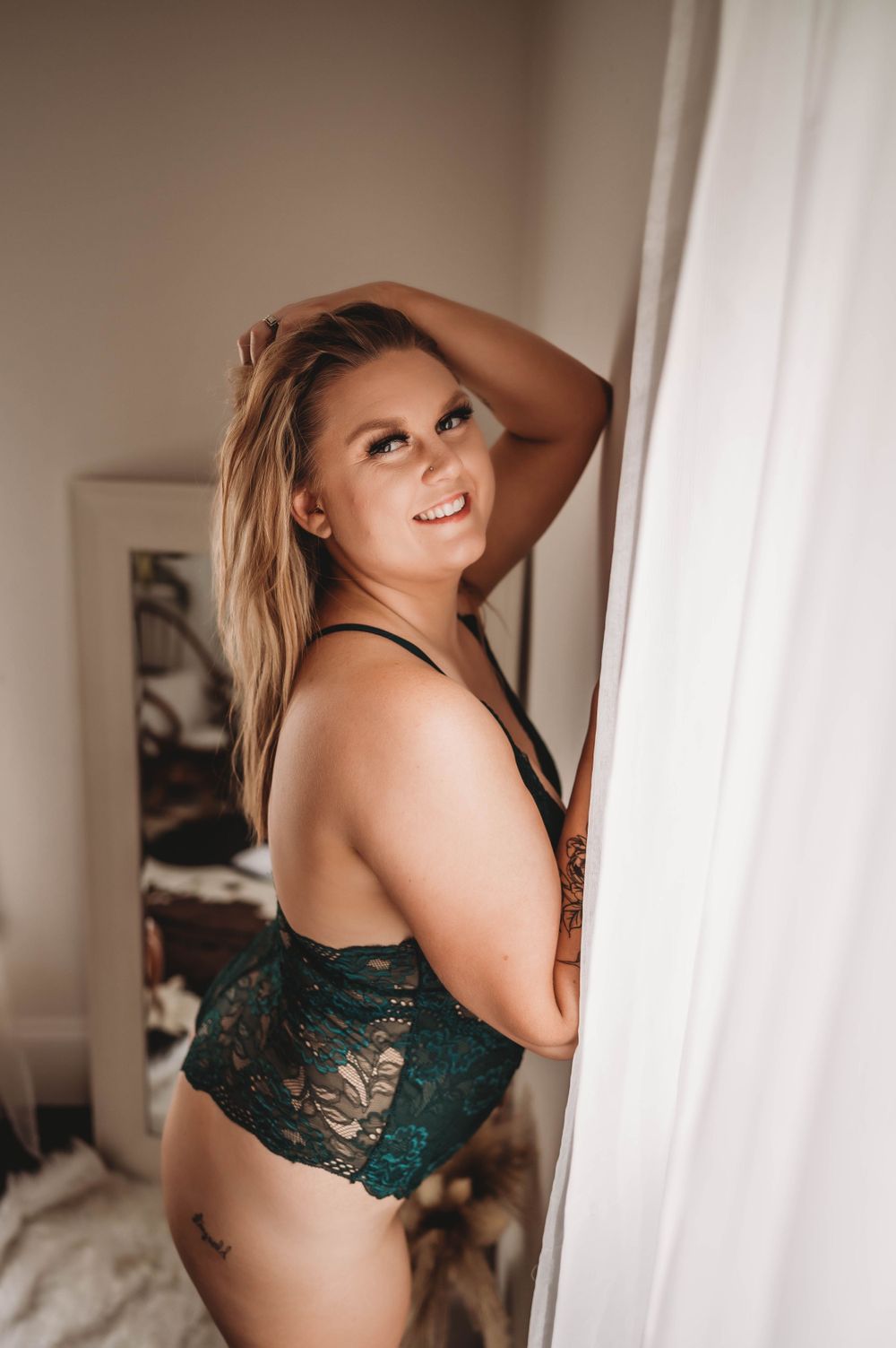 A woman in emerald lace lingerie poses by a window in natural light.