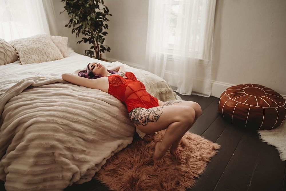 A woman in red loungewear relaxes on a bed with fur rug and leather pouf decor.