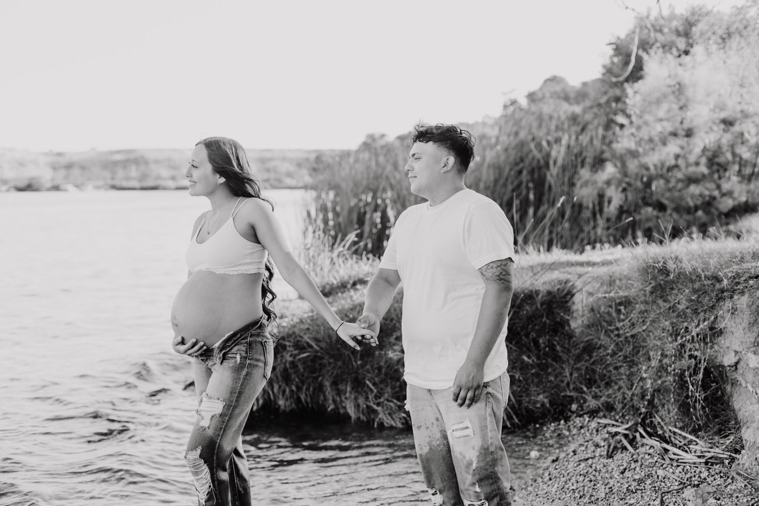 Black and white photo of a couple standing by a lake during a maternity shoot.
