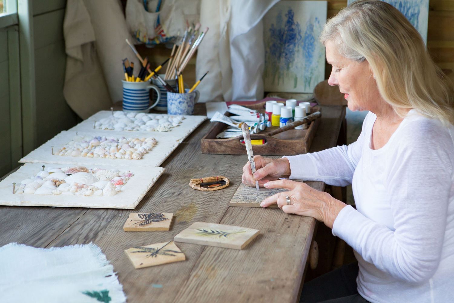 Artist working on detailed watercolor paintings at a rustic wooden desk with art supplies.