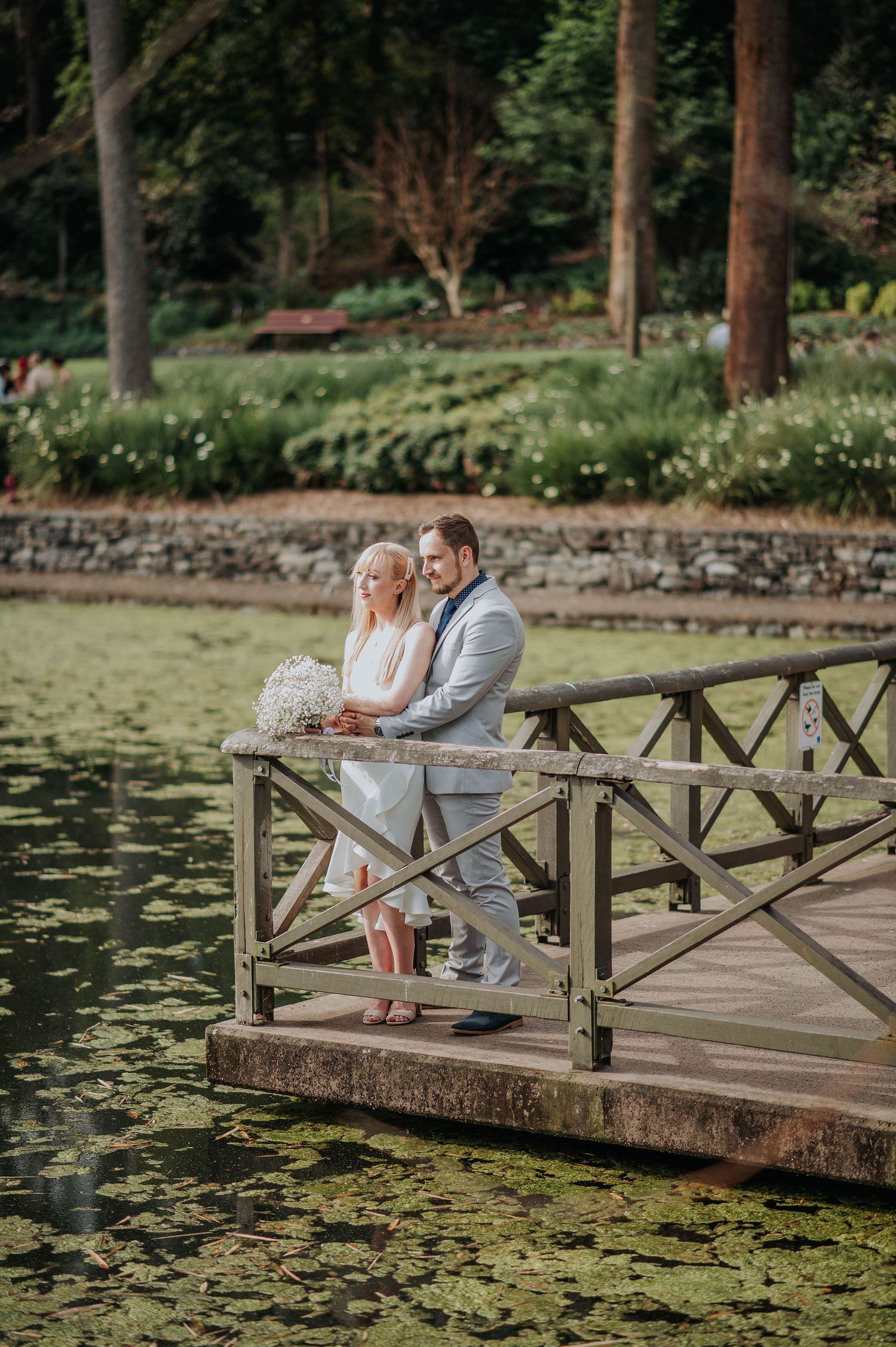 Newlyweds embrace on wooden dock overlooking lily pond in serene garden setting.