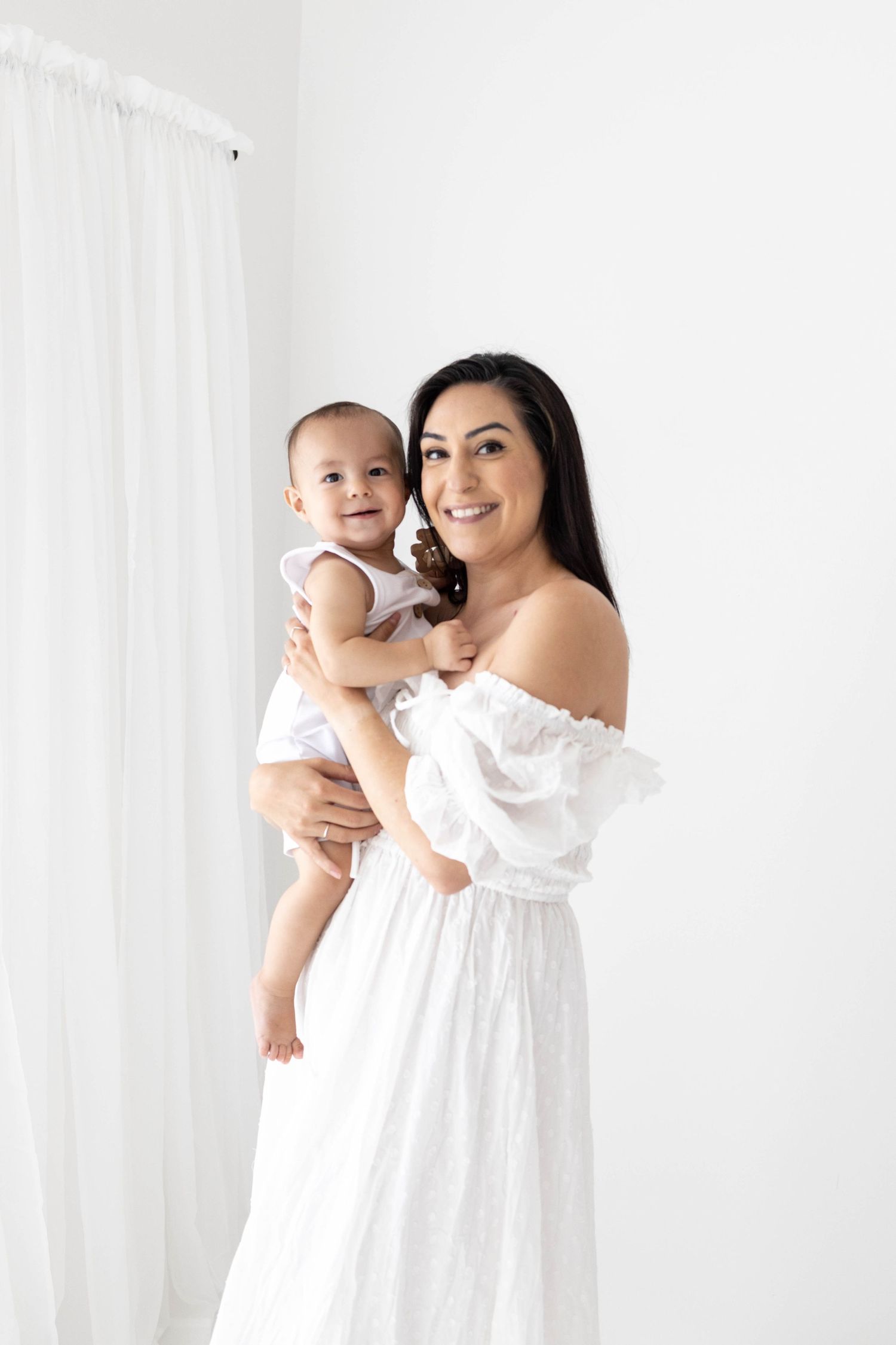 A mother in a white dress holds and cuddles with her baby against a light background.