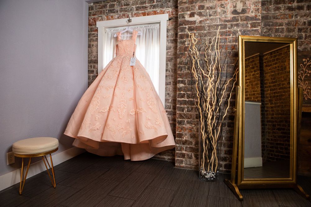 A blush pink ballgown hangs elegantly against an exposed brick wall next to a gold-framed mirror and decorative branches.