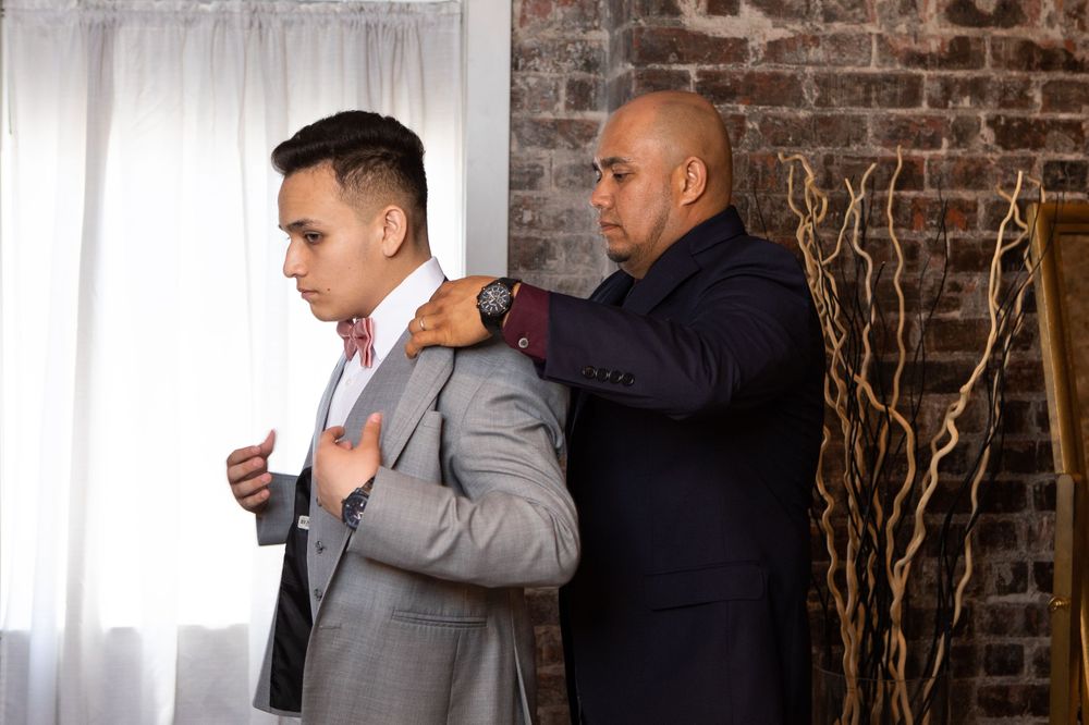 A groom getting help with his light gray suit jacket during wedding preparations.