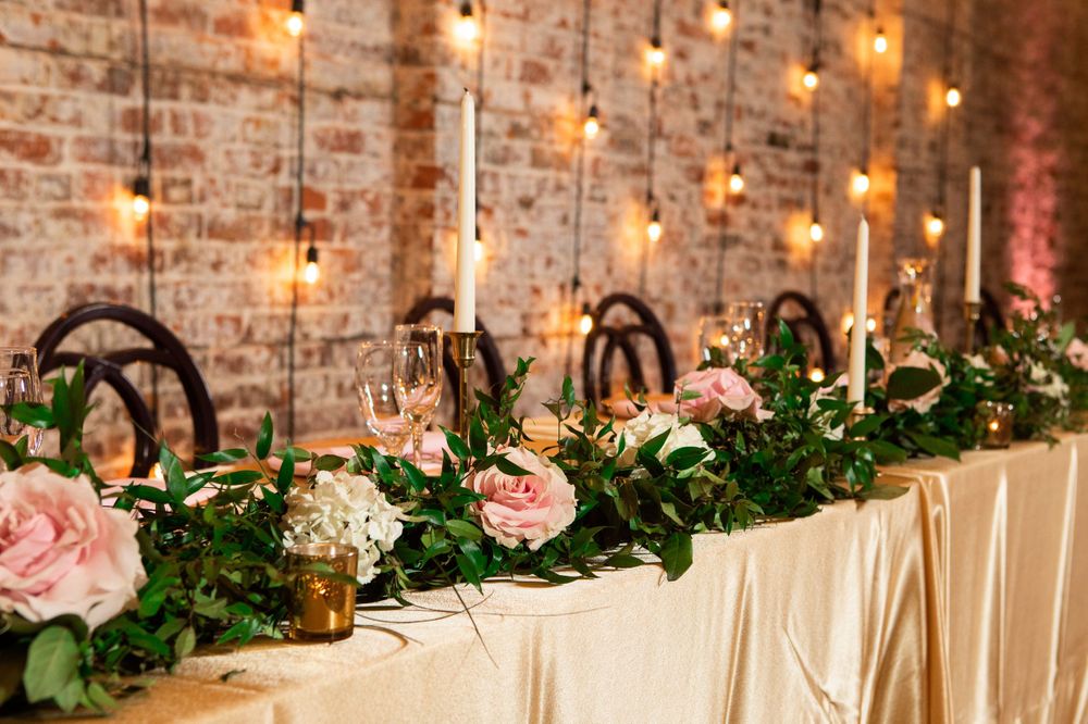 Wedding table decorated with pink roses, greenery, and gold accents along a white tablecloth.