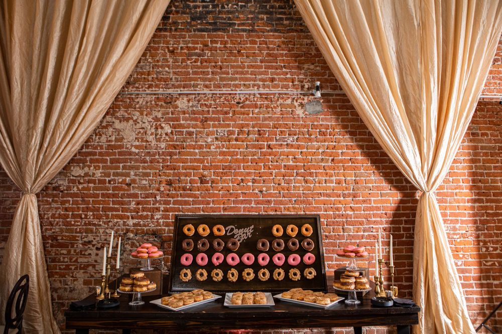 Dessert table with donuts display against exposed brick wall with champagne colored drapes.