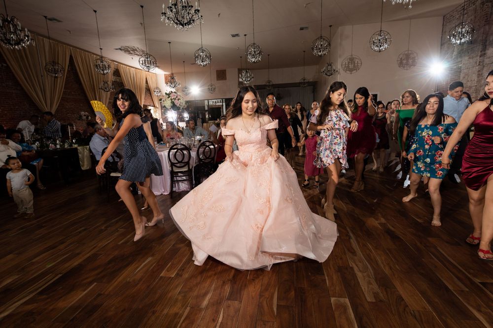 A pink ballgown twirls on a wooden dance floor while party guests dance in colorful dresses.
