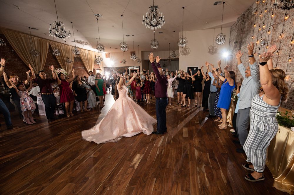 A celebration shows dancers in colorful attire surrounding a flowing pink gown on a hardwood floor.