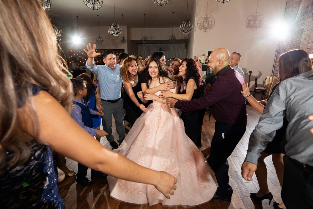 Party guests dance and celebrate surrounding someone in a pink ballgown at an indoor venue.